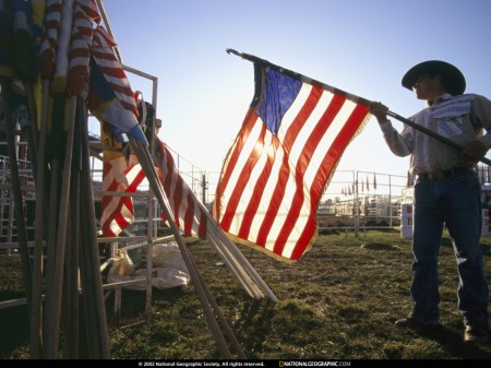 American Flag, Spencer, Iowa, 1996 – caption from the National Geographic Society: A man rolls up U.S. flags at the end of the Clay County Fair in Spencer, Iowa. “Although the population of Spencer is only about 12,000, the fair draws some 300,000 visitors. Once a year, rising from the endless flatness of the Iowa countryside, a crowd forms—to stroll, to hear big country music acts like the Statler Brothers, to sell a grand champion boar, to buy a new silo.” (Photographed on assignment for, but not published in, “County Fairs,” October 1997, National Geographic magazine) Photograph by Randy Olson; copyright National Geographic Society